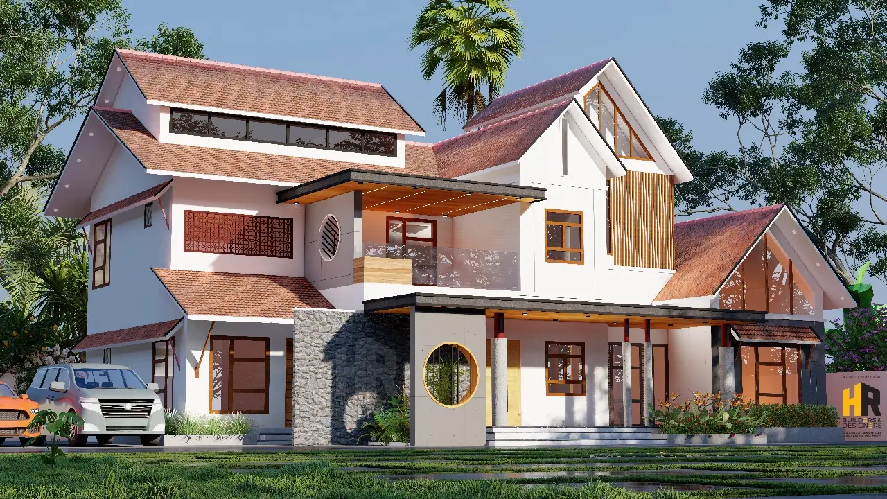 The house is white with a complex, reddish-brown tile roof that has multiple gables and peaks. The building has a prominent second-floor balcony with a glass railing and a wooden ceiling, supported by a mix of circular and square pillars.designed by hr buliders kunnamkulam